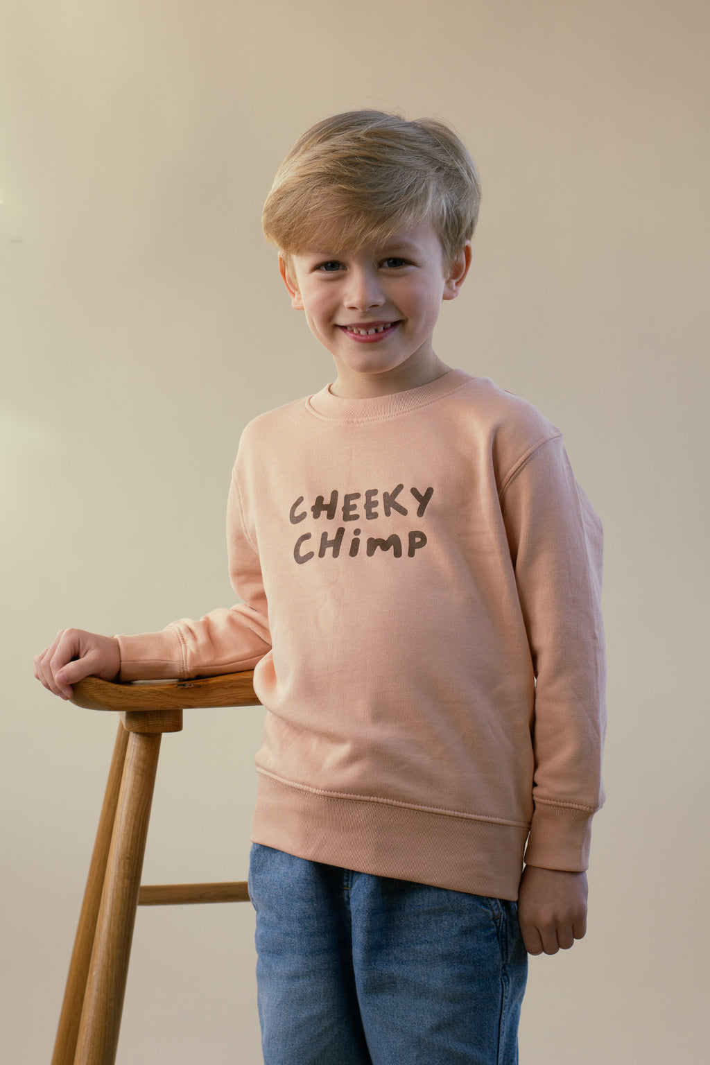 Child wearing a peach sweatshirt with 'Cheeky Chimp' text, standing bu a wooden stool against a plain background.