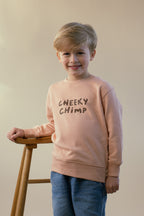 Child wearing a peach sweatshirt with 'Cheeky Chimp' text, standing bu a wooden stool against a plain background.