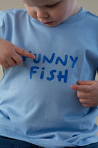 Child wearing a blue t-shirt with 'FUNNY FISH' text, pointing at it