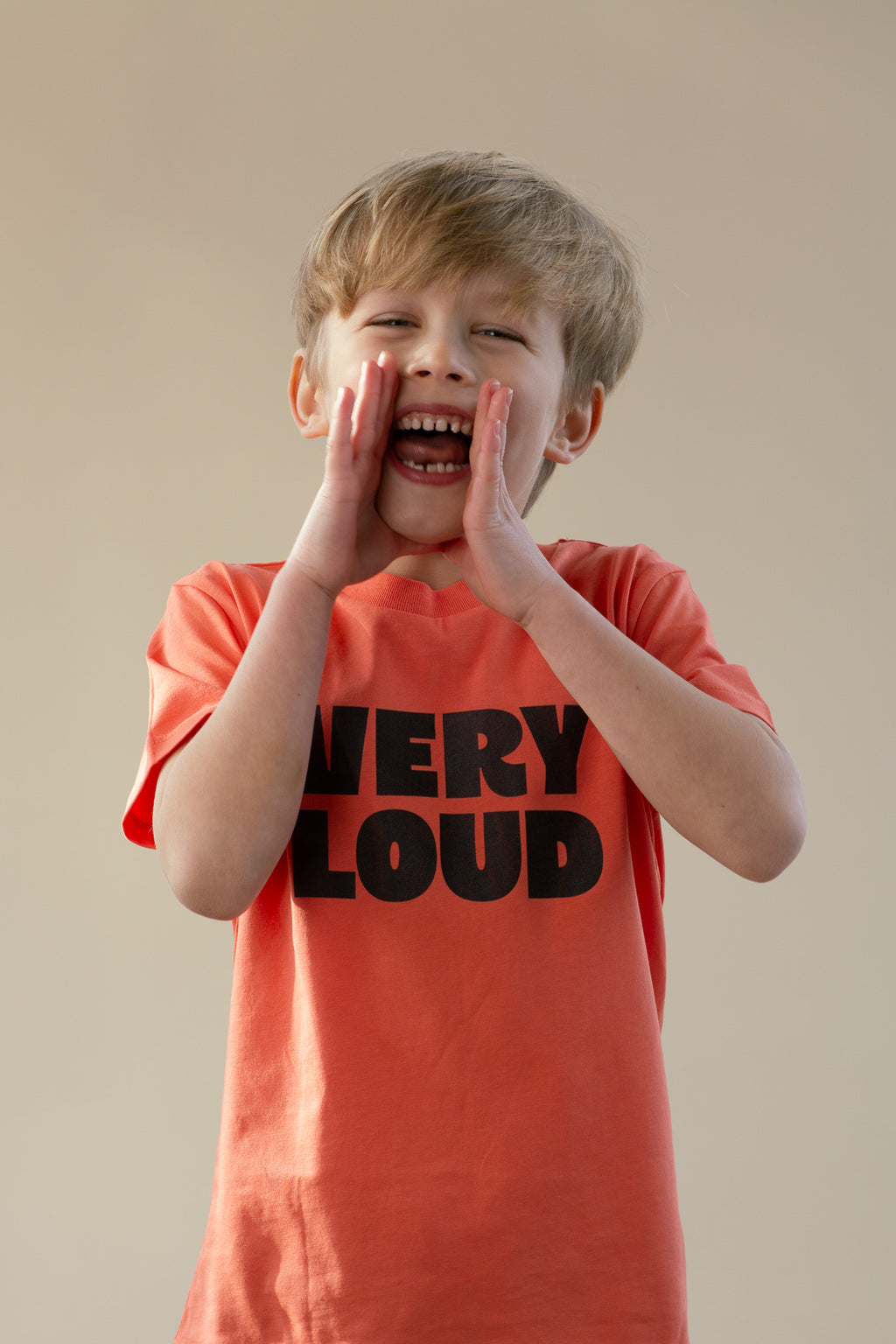 Child wearing a 'VERY LOUD' t-shirt, shouting with hands over mouth against a plain background
