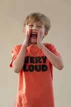 Child wearing a 'VERY LOUD' t-shirt, shouting with hands over mouth against a plain background
