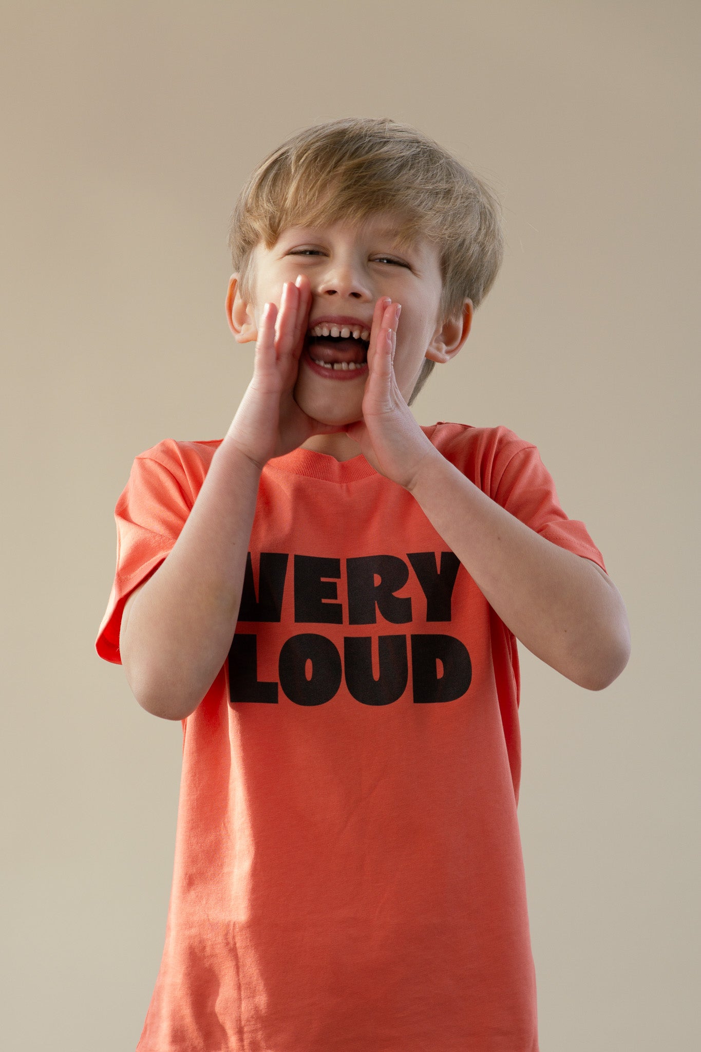 Child wearing a 'VERY LOUD' t-shirt, shouting with hands over mouth against a plain background