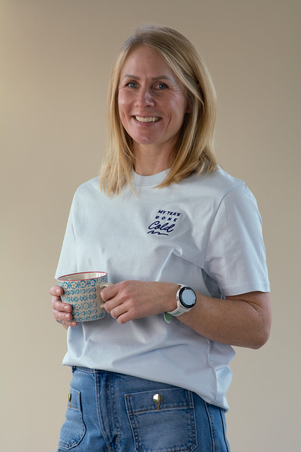Woman holding a mug and wearing a blue t-shirt with a logo against a beige background