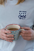 Person wearing a blue t-shirt with 'My Tea's Gone Cold' text, holding a patterned mug