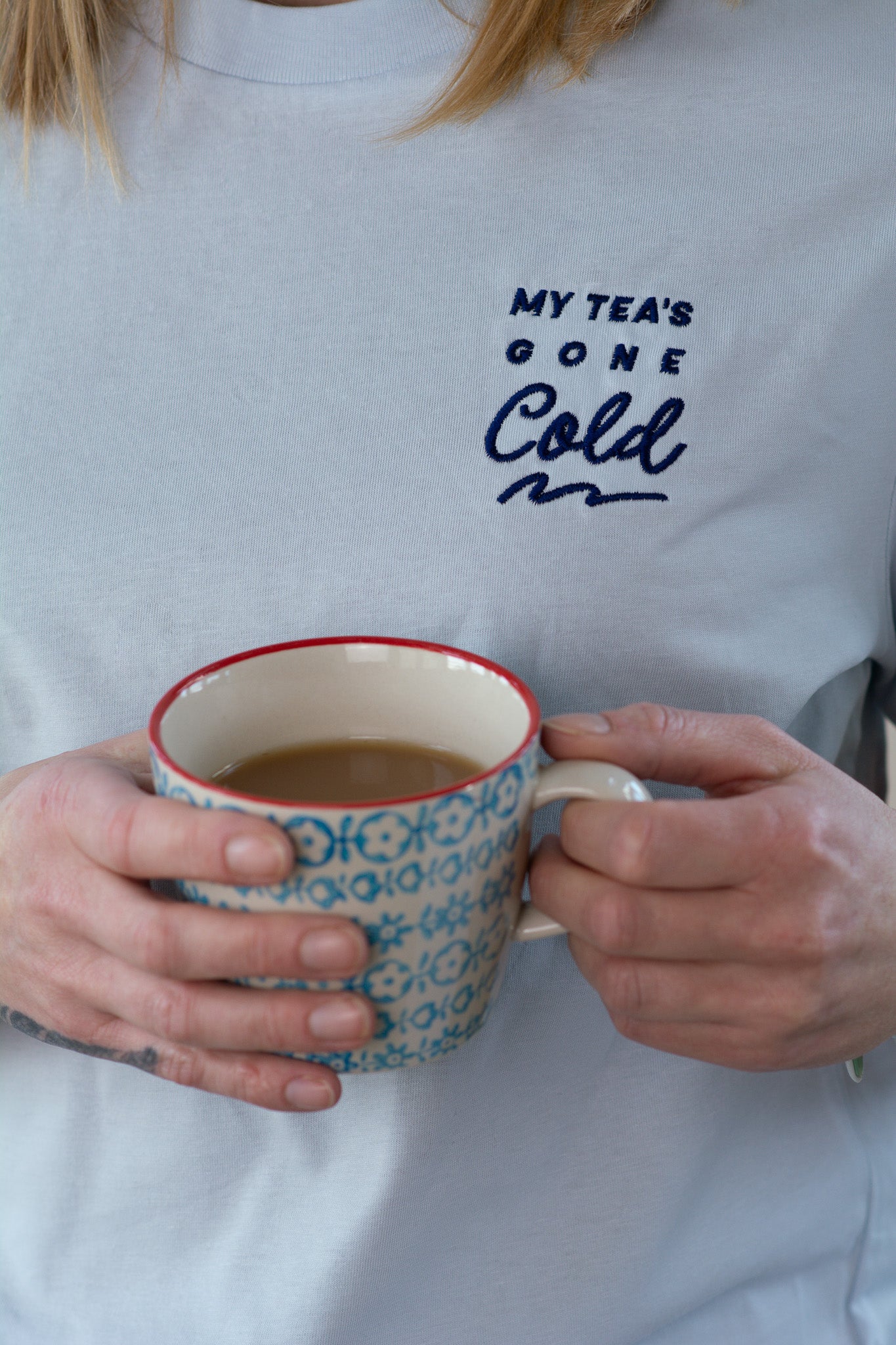 Person wearing a blue t-shirt with 'My Tea's Gone Cold' text, holding a patterned mug