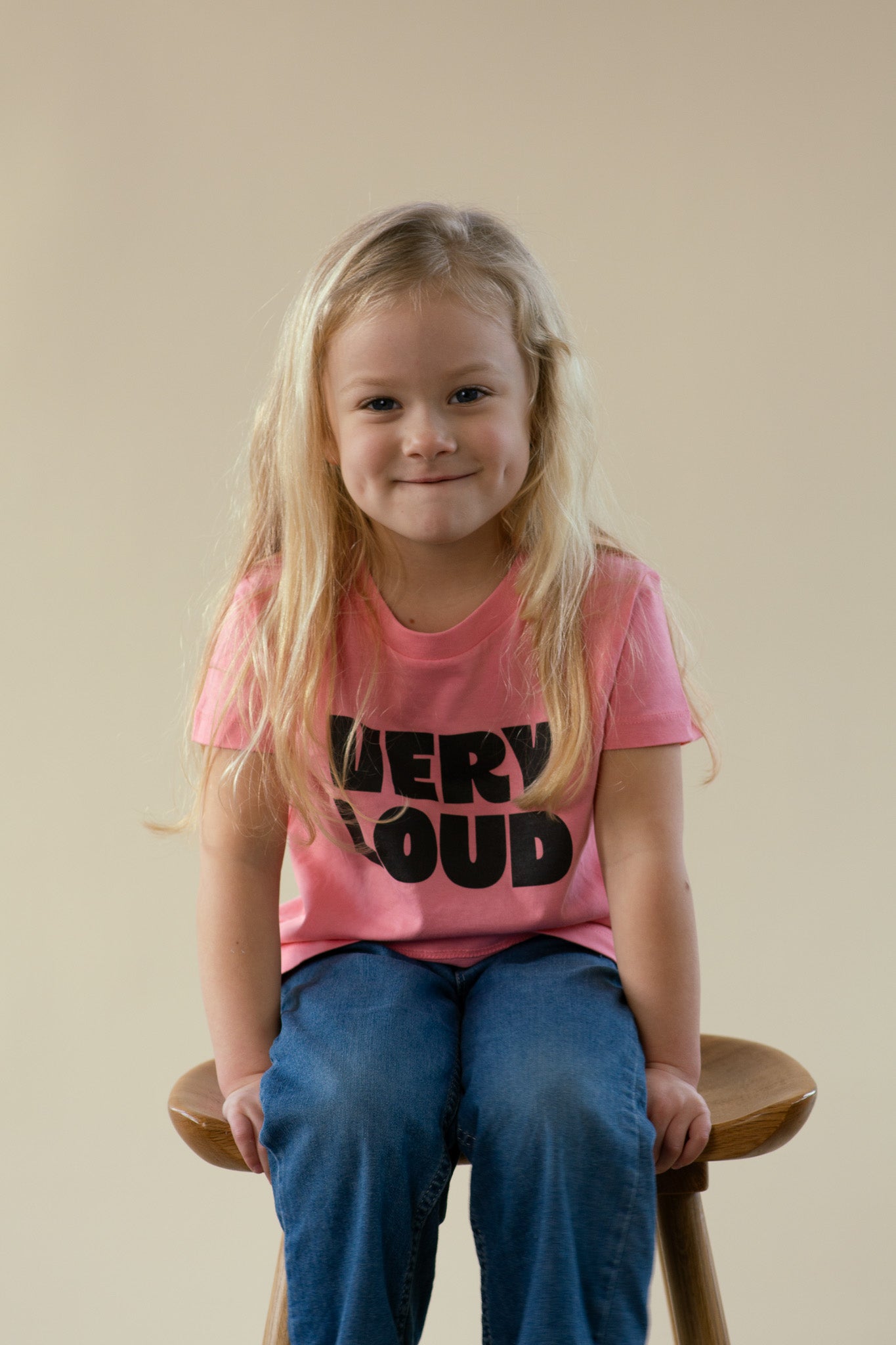 Child wearing a pink shirt with 'VERY LOUD' text, sitting on a wooden stool against a beige background