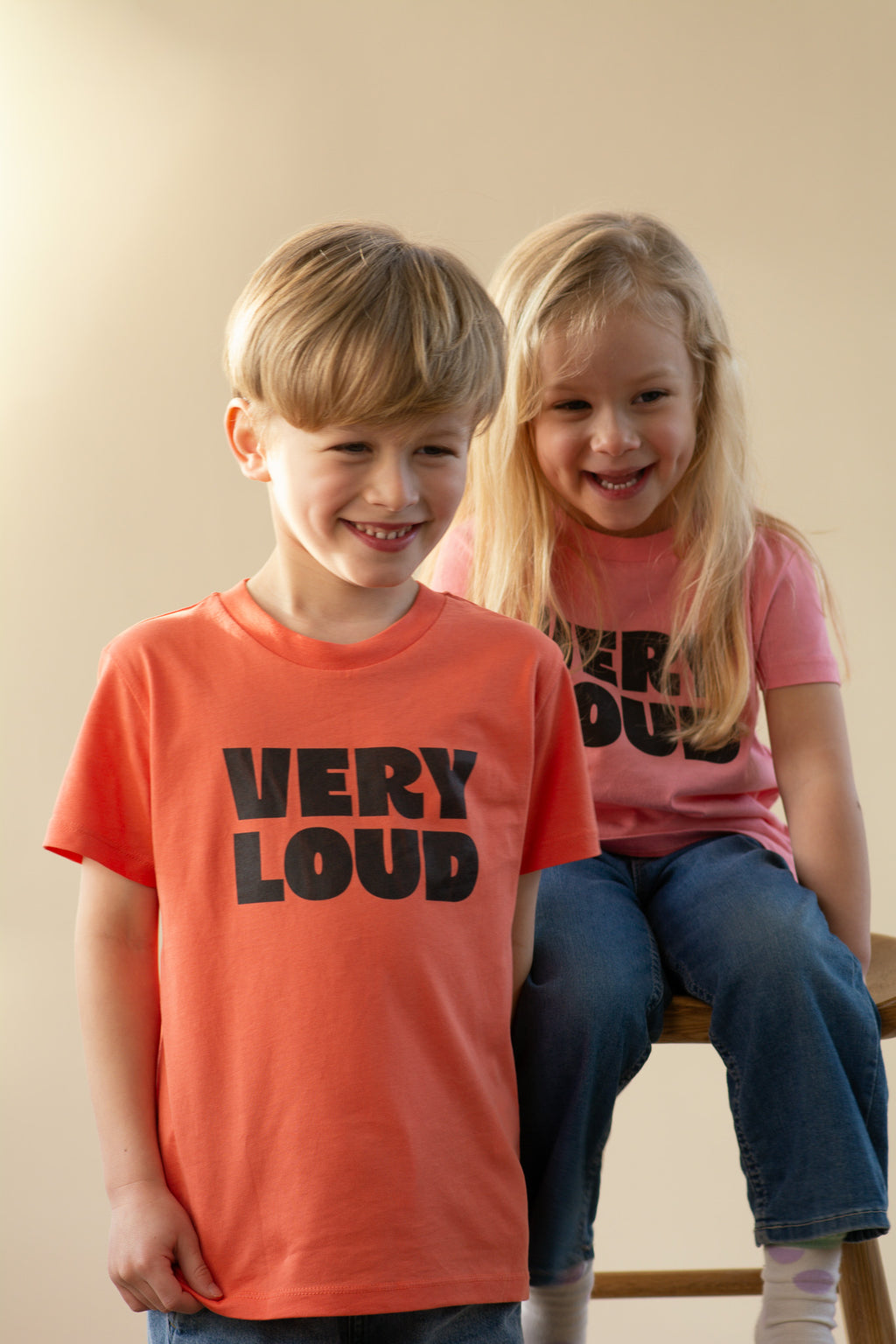 Two children wearing 'VERY LOUD' t-shirts sitting on a stool against a plain background