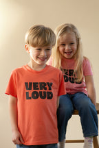 Two children wearing 'VERY LOUD' t-shirts sitting on a stool against a plain background