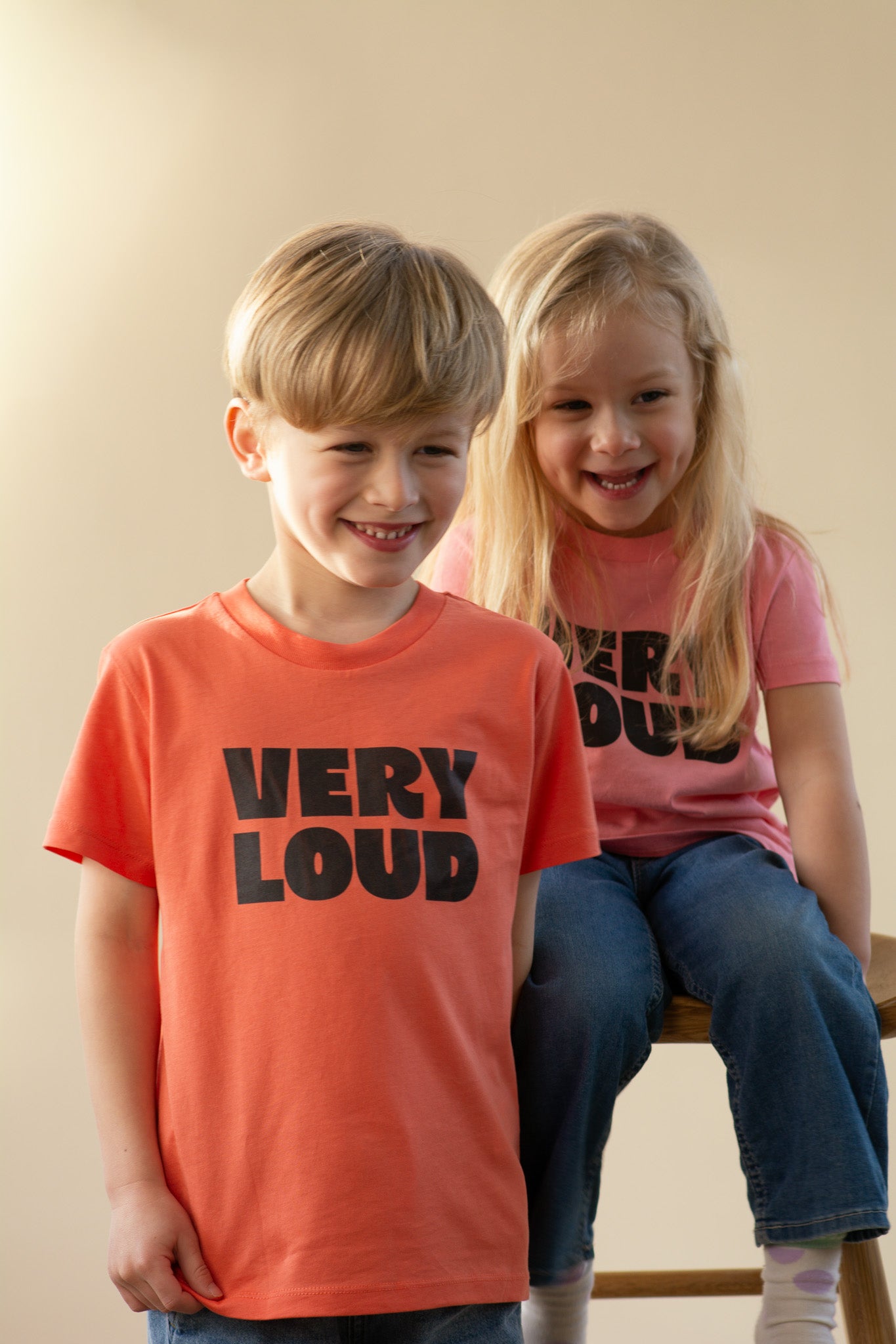Two children wearing 'VERY LOUD' t-shirts sitting on a stool against a plain background
