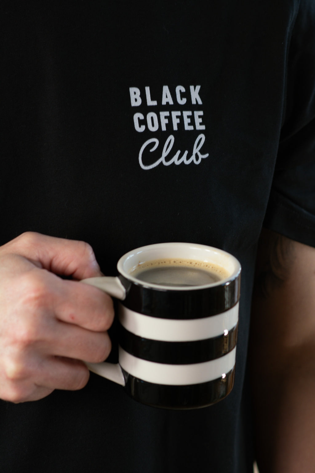 Person holding a black and white striped mug with 'Black Coffee Club' text on a black shirt.