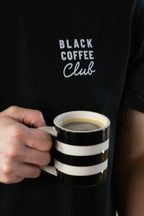 Person holding a black and white striped mug with 'Black Coffee Club' text on a black shirt.