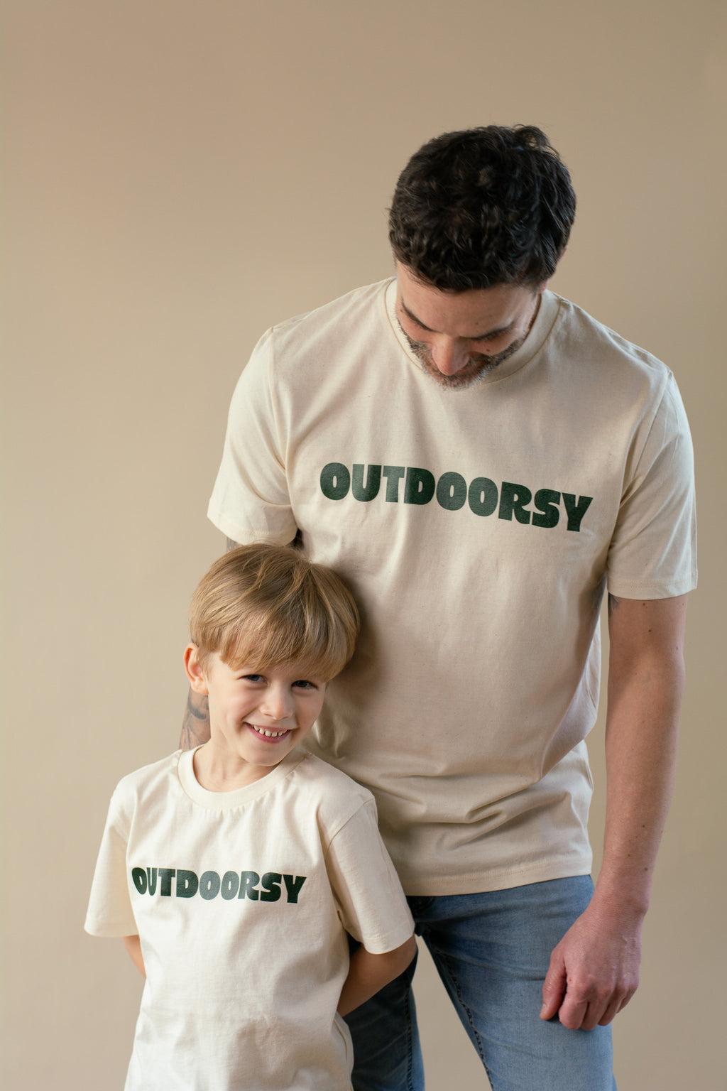 Man and child wearing 'OUTDOORSY' t-shirts against a beige background