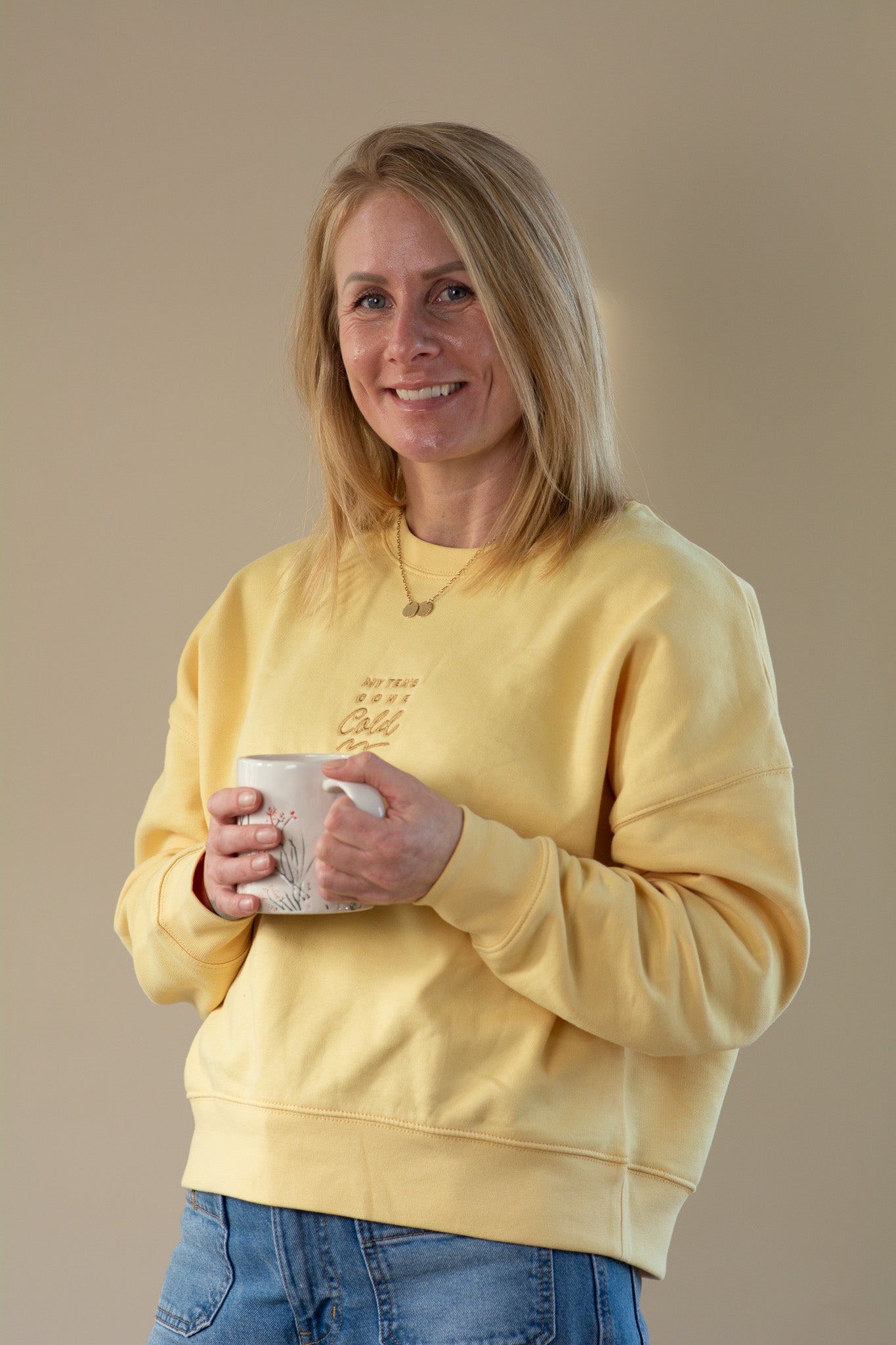 Woman wearing a yellow sweatshirt holding a mug against a beige background