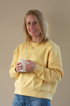 Woman wearing a yellow sweatshirt holding a mug against a beige background