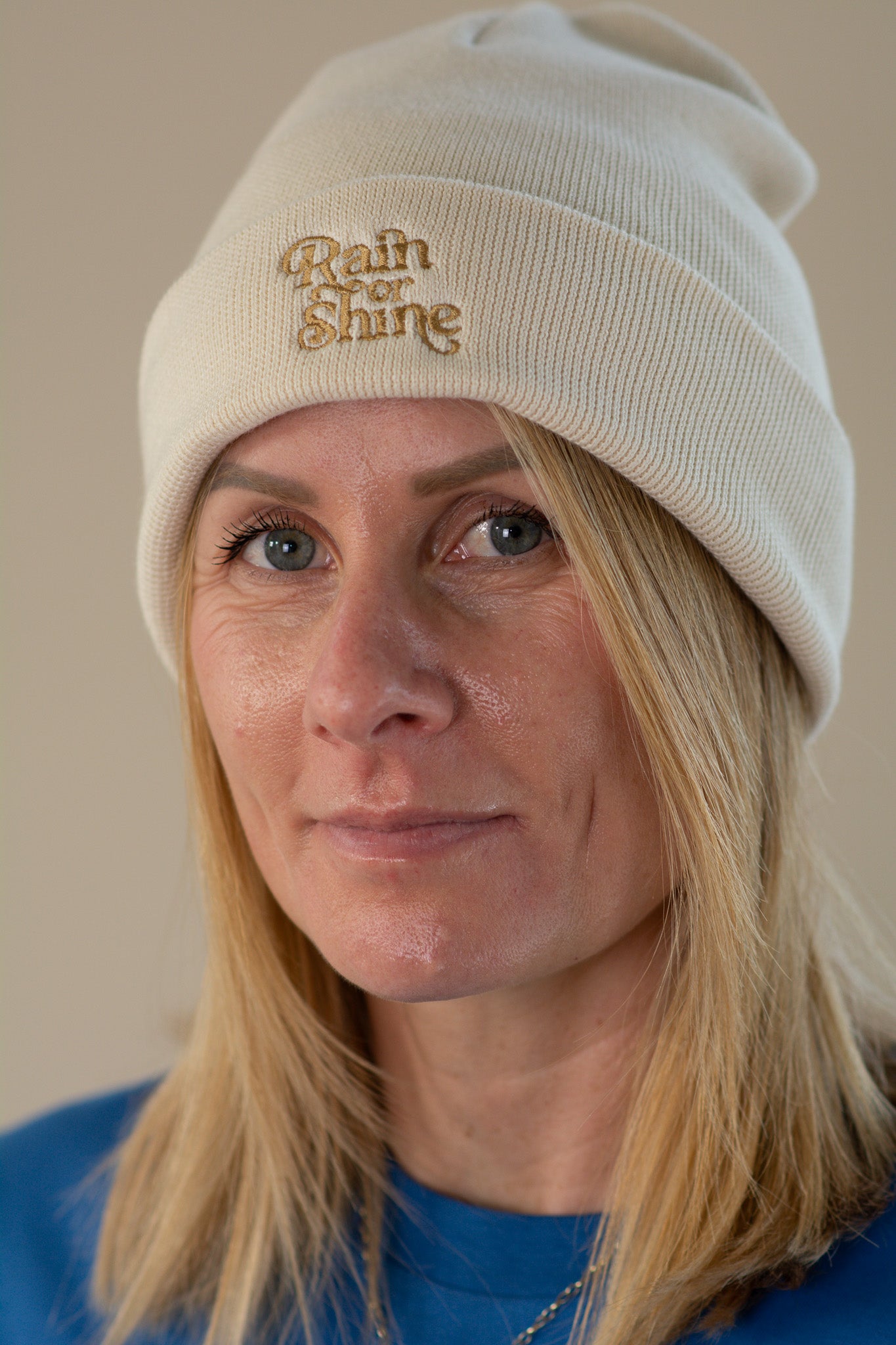 Person wearing a beige beanie with 'Rain or Shine' text against a plain background