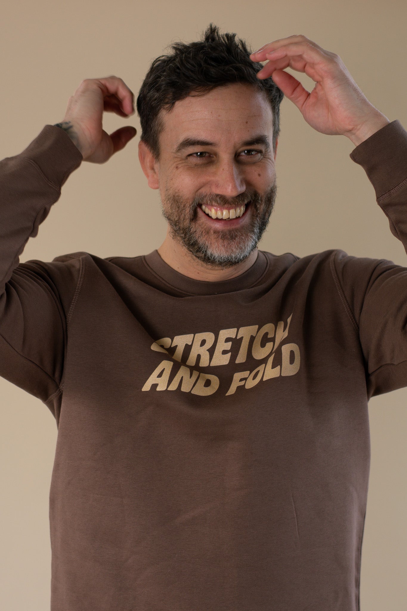 Man wearing a brown sweatshirt with 'STRETCH AND FOLD' text, posing with hands on head against a beige background.