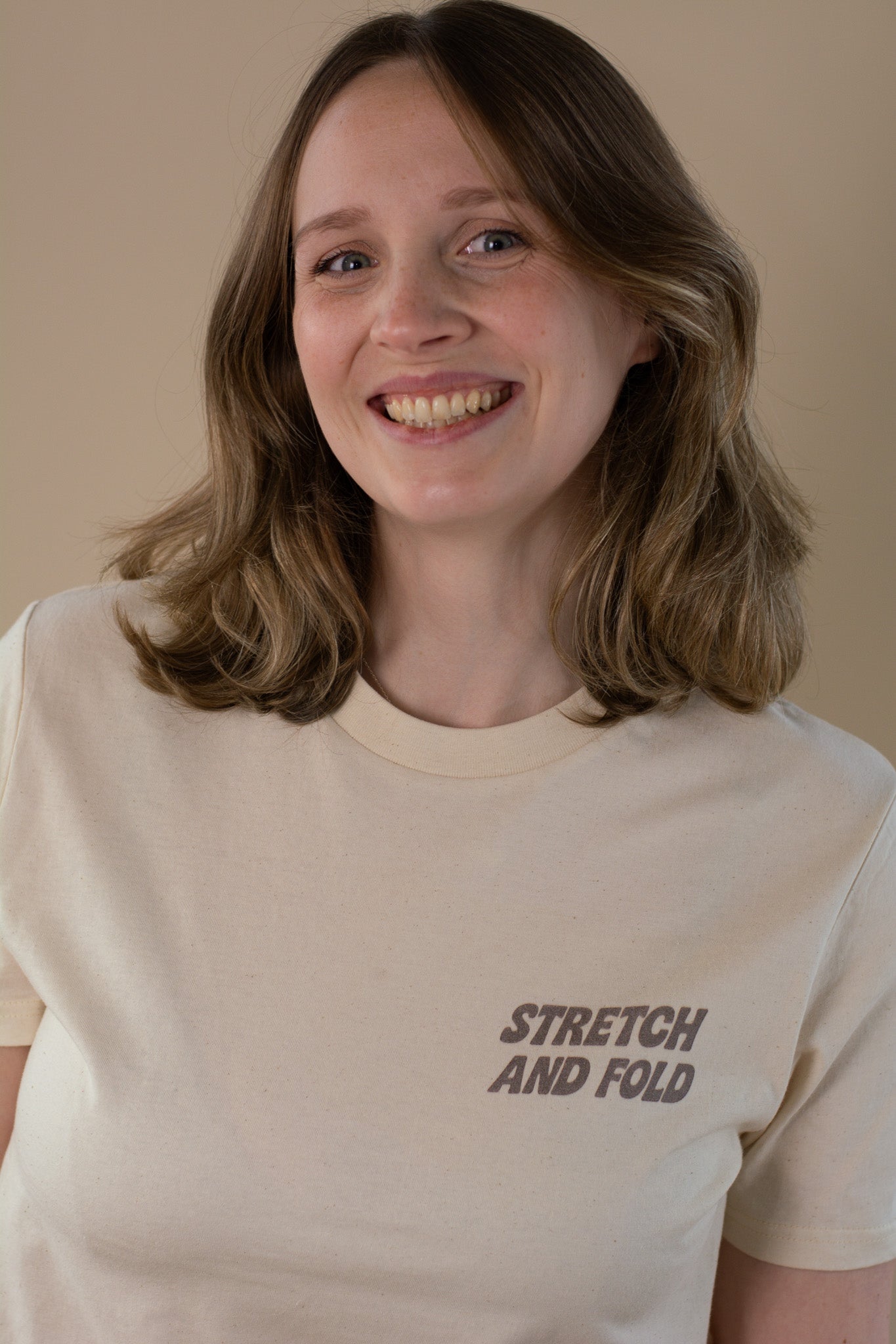 Person wearing a beige t-shirt with 'STRETCH AND FOLD' text on a plain background