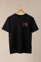 Black t-shirt with orange logo on a wooden hanger against a beige background