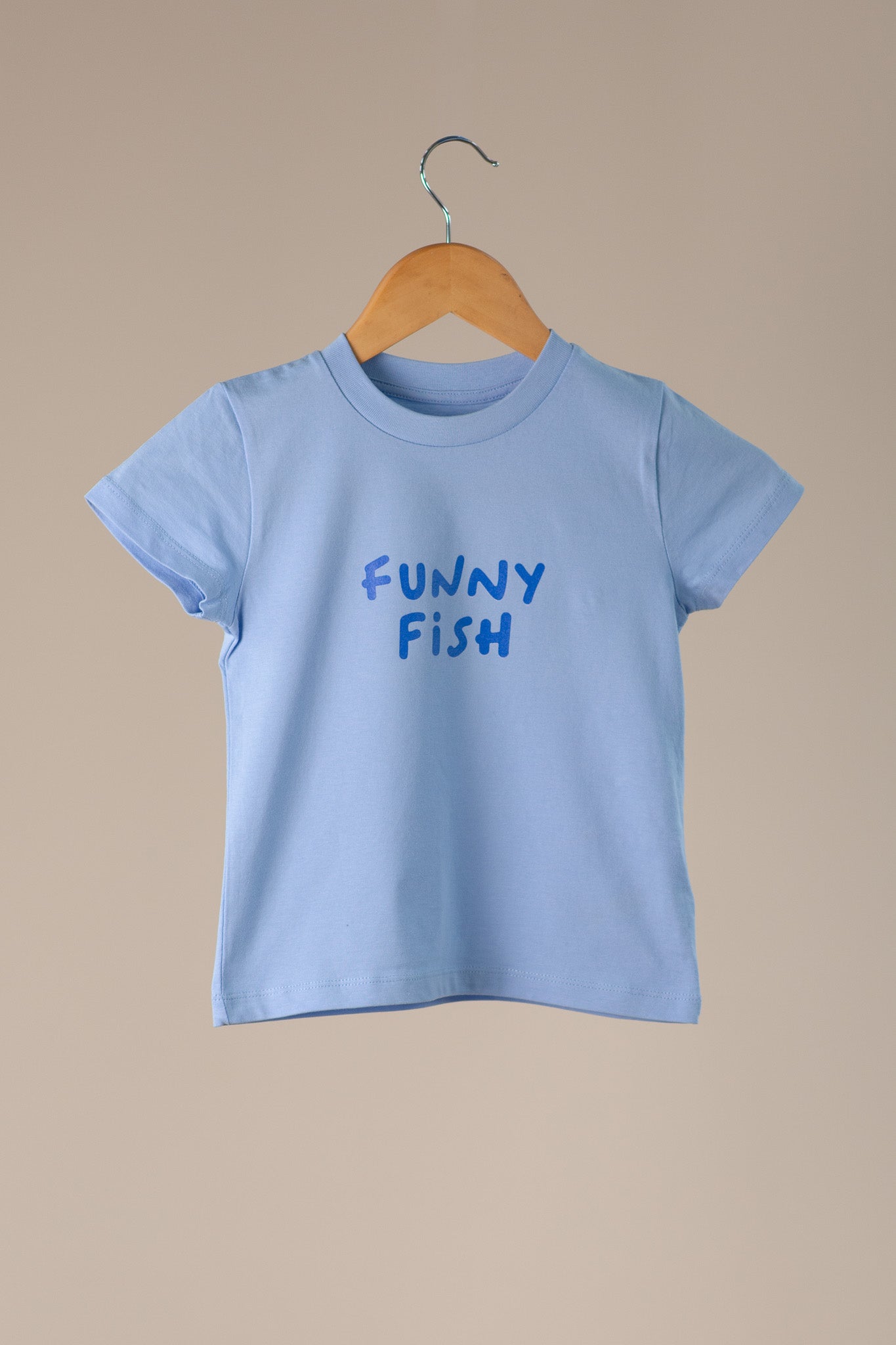 Light blue t-shirt with 'FUNNY FISH' text on a wooden hanger against a beige background