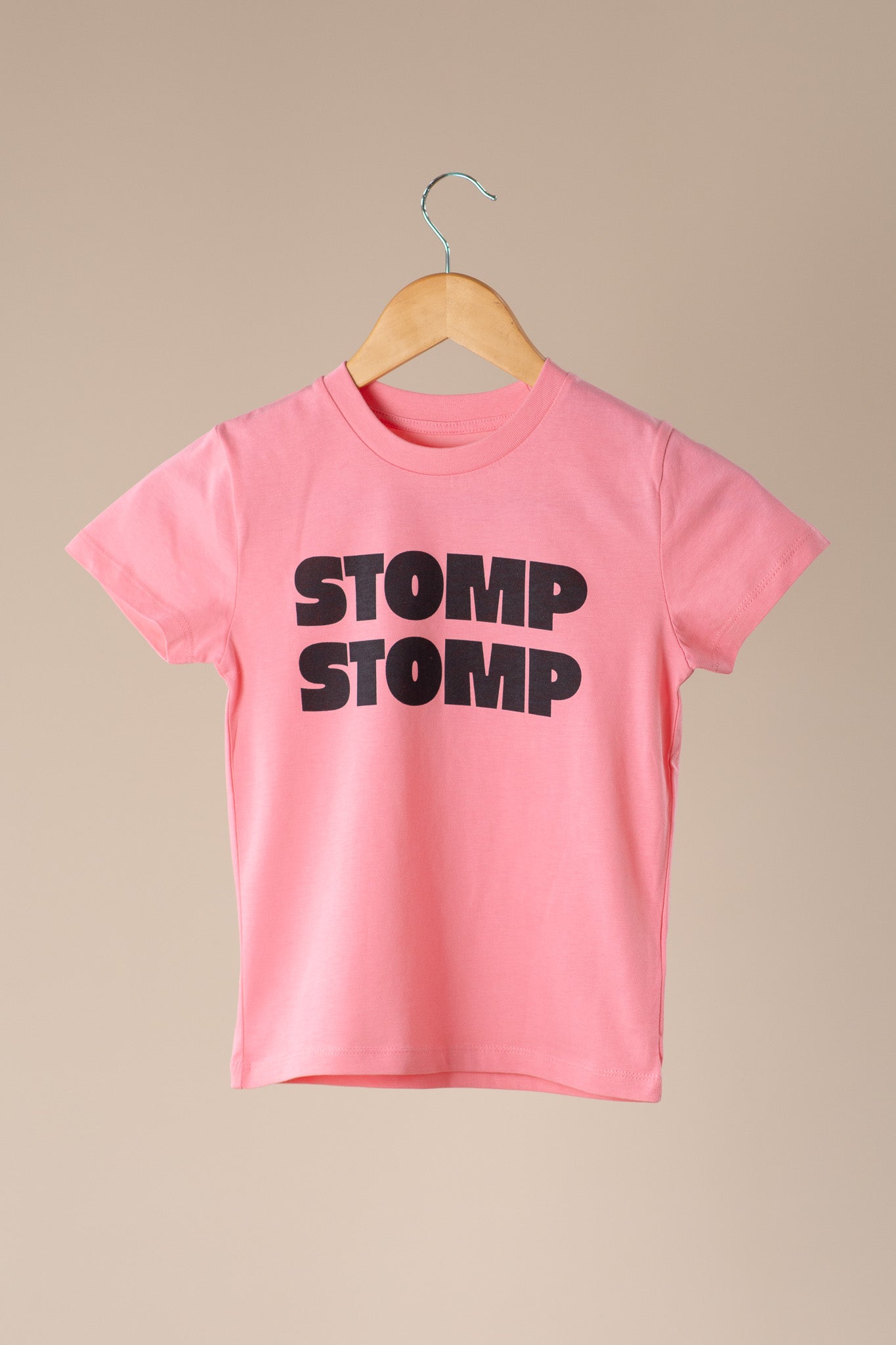Pink t-shirt with 'STOMP STOMP' text on a wooden hanger against a beige background