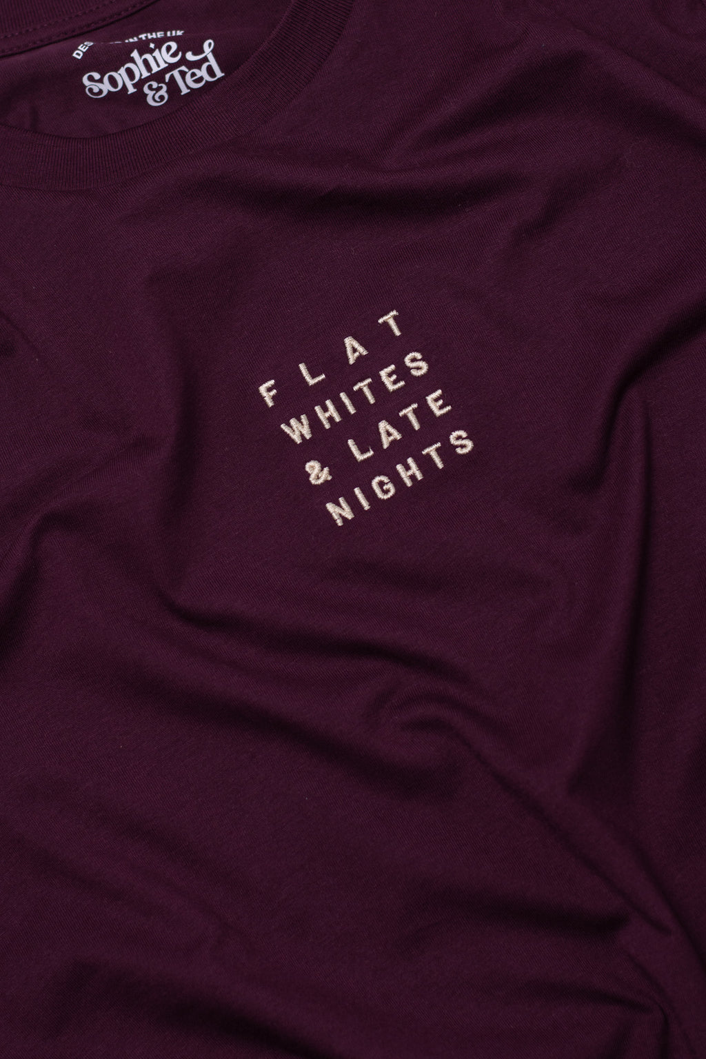 Maroon t-shirt with text 'FLAT WHITES & LATE NIGHTS' on a close-up of fabric texture
