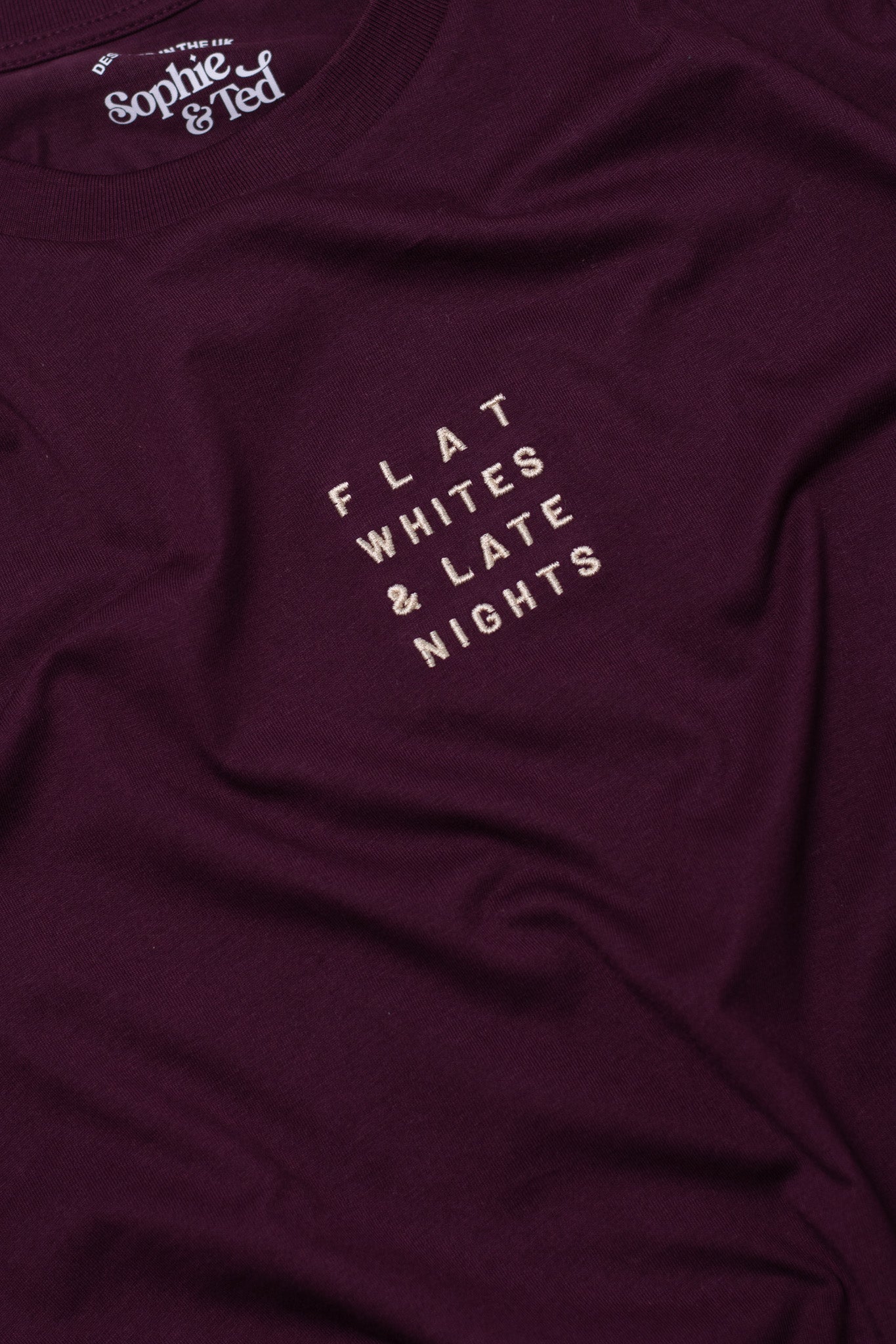 Maroon t-shirt with text 'FLAT WHITES & LATE NIGHTS' on a close-up of fabric texture