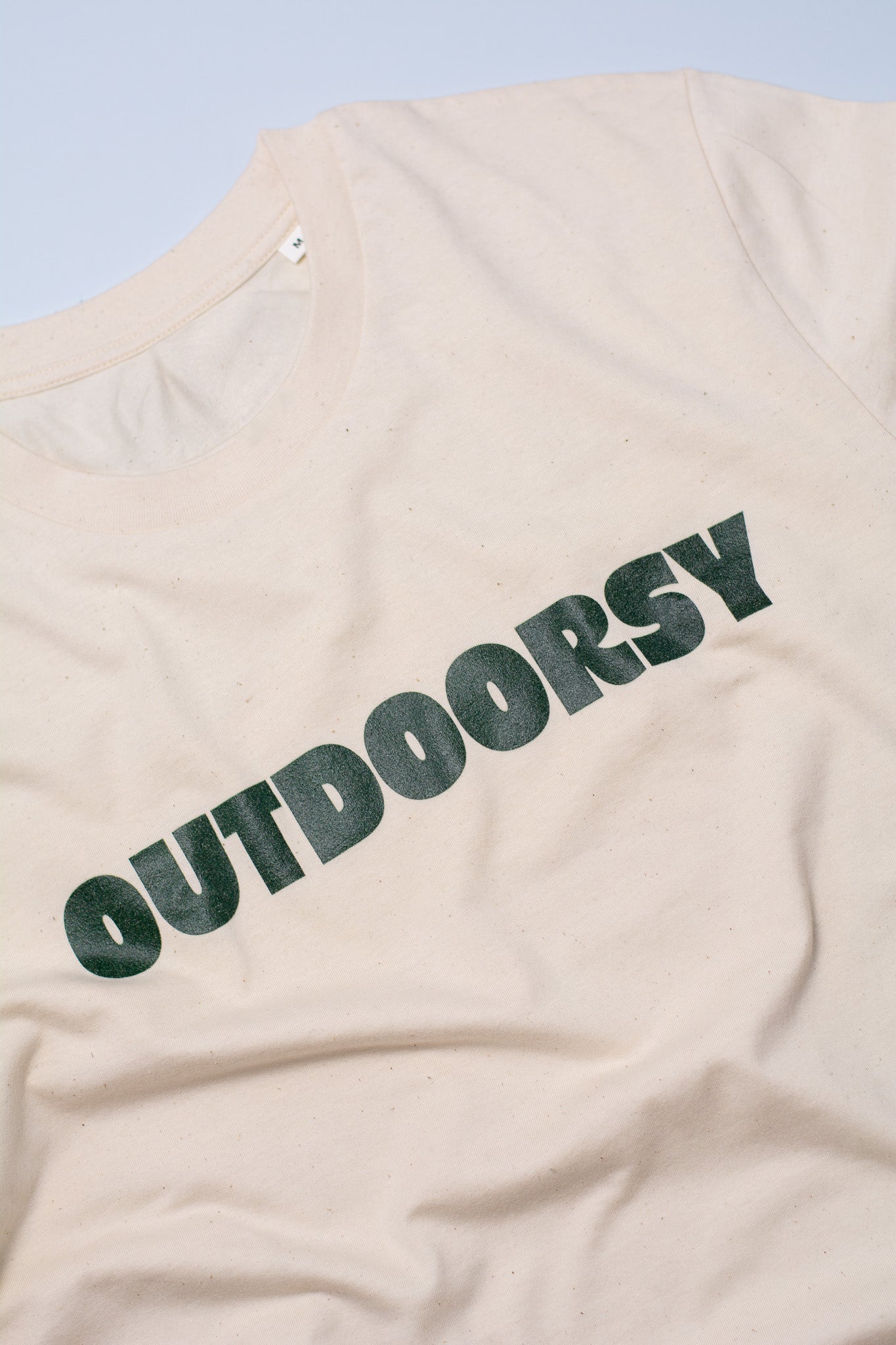 Beige t-shirt with 'OUTDOORSY' text on a light blue background