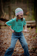 Child wearing a teal shirt with 'SILLY GOOSE' text, blue jeans, and striped hat outdoors.