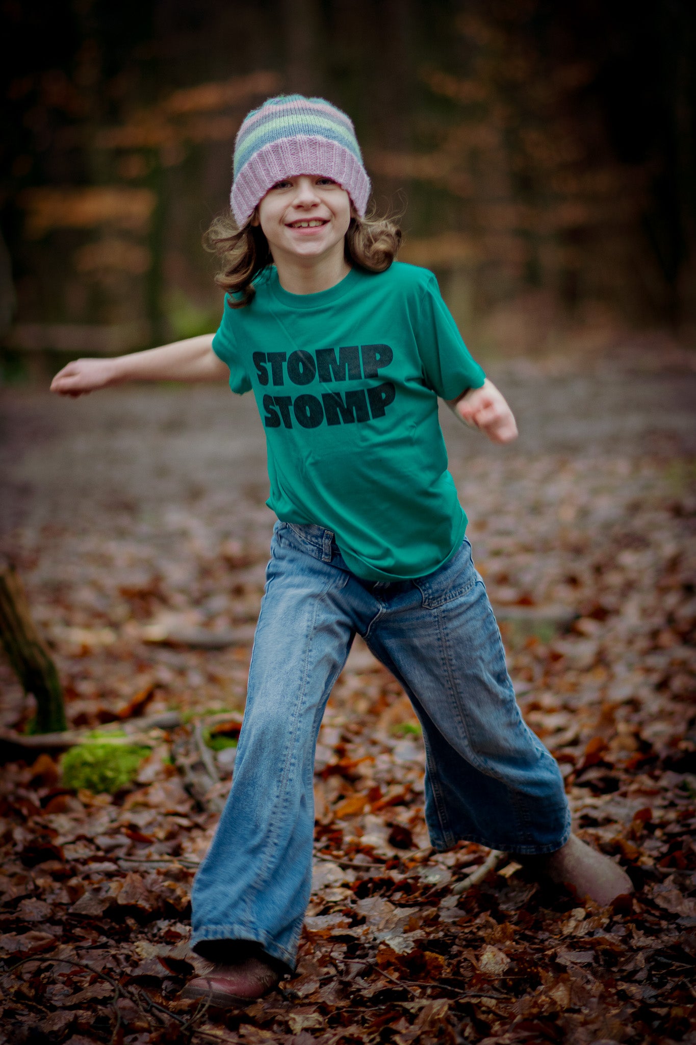 Child wearing a green t-shirt with 'STOMP' text, blue jeans, and a striped hat in a forest setting.