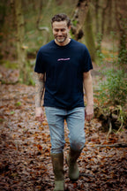 Man walking in a forest wearing a navy t-shirt and green boots.