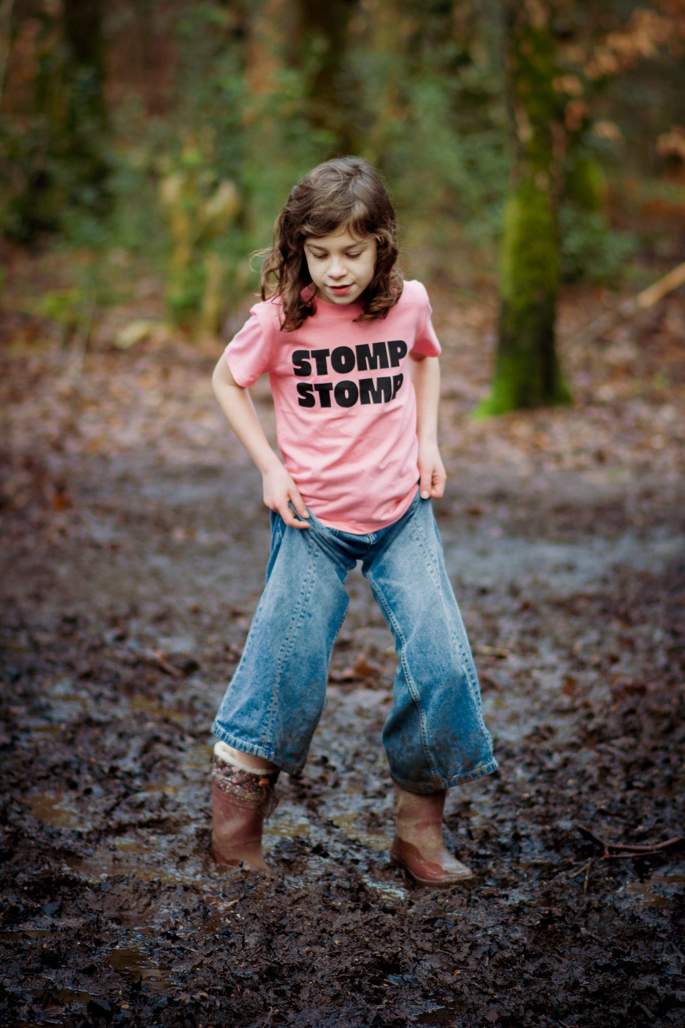 Child wearing a pink shirt with 'STOMP STOMP' text, blue jeans, and brown boots standing in a muddy forest floor.