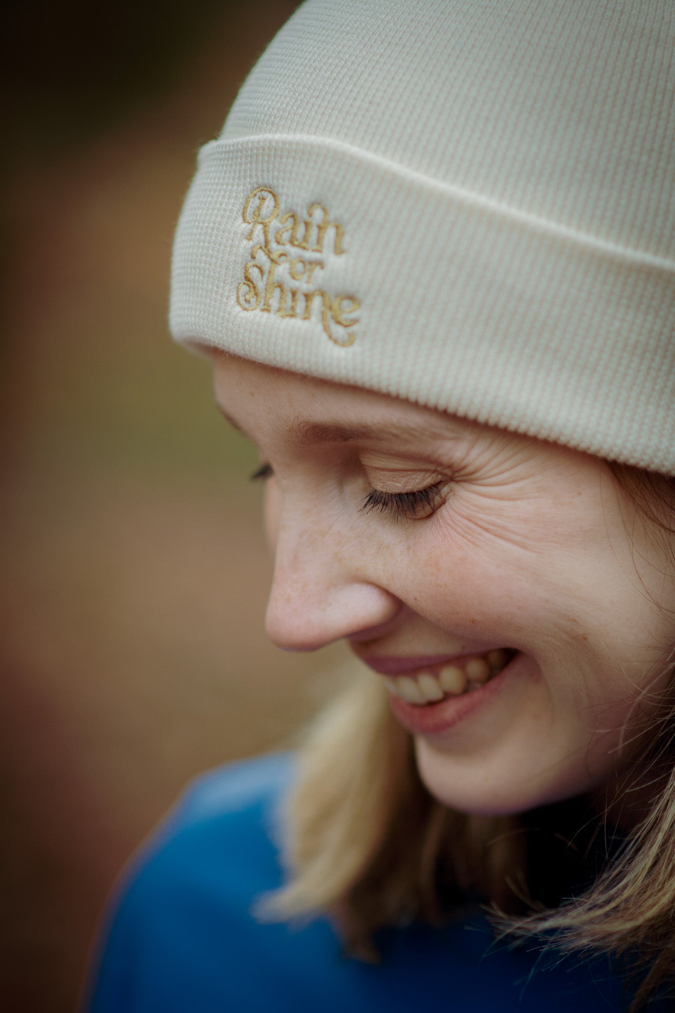 Person wearing a beige beanie with 'Rain or Shine' embroidery, smiling outdoors.