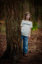 Child wearing a 'Outdoorsy' shirt standing next to a tree in a forest