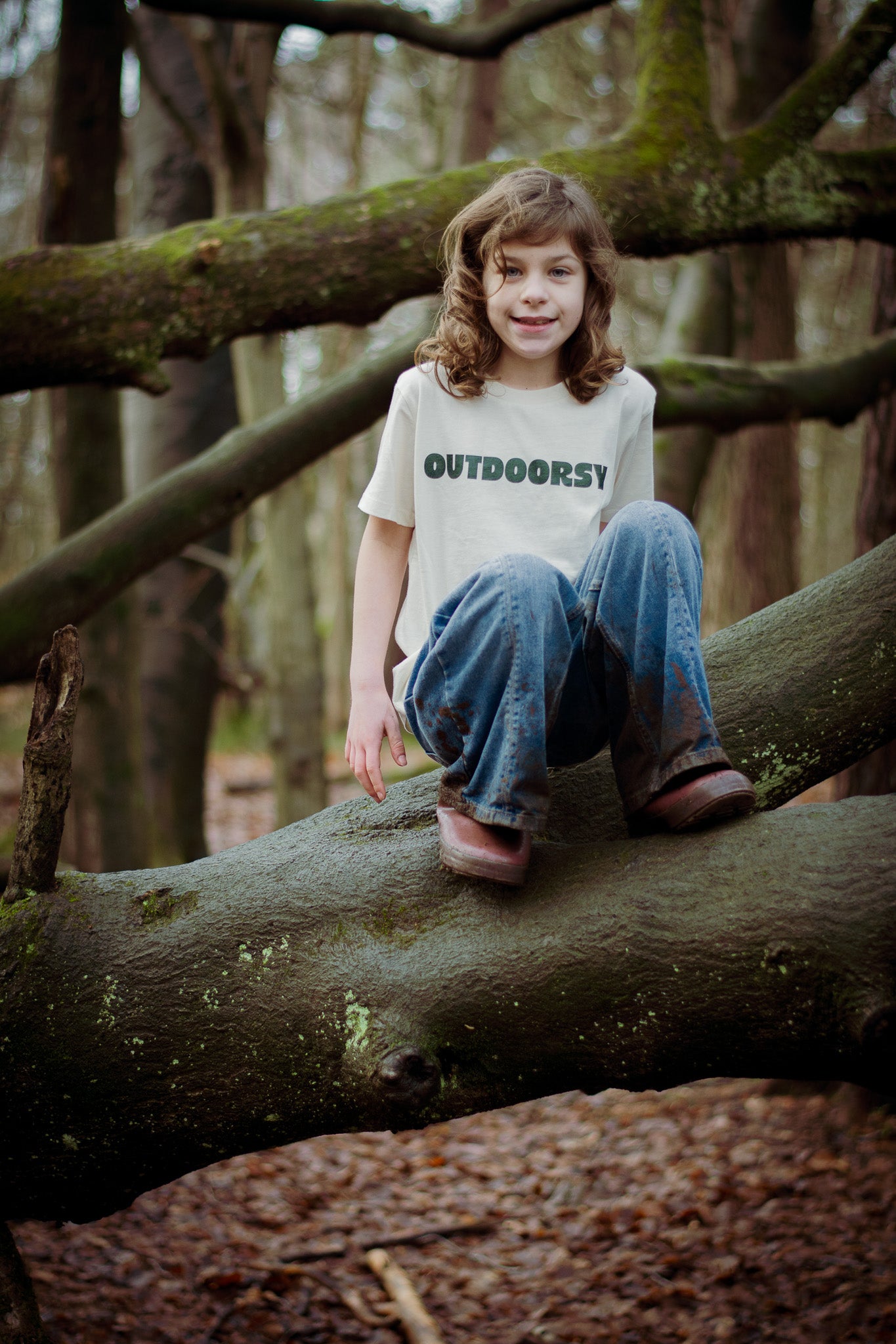 Child sitting on a tree branch in a forest wearing a 'OUTDOORSY' t-shirt