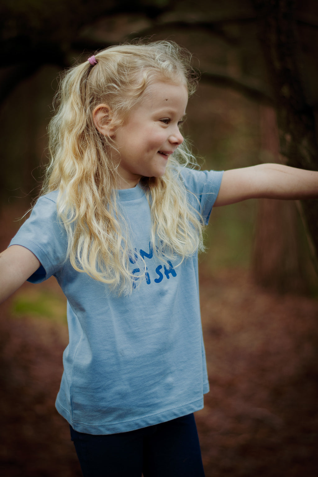 Young girl with blonde hair wearing a blue t-shirt with text, standing outdoors.