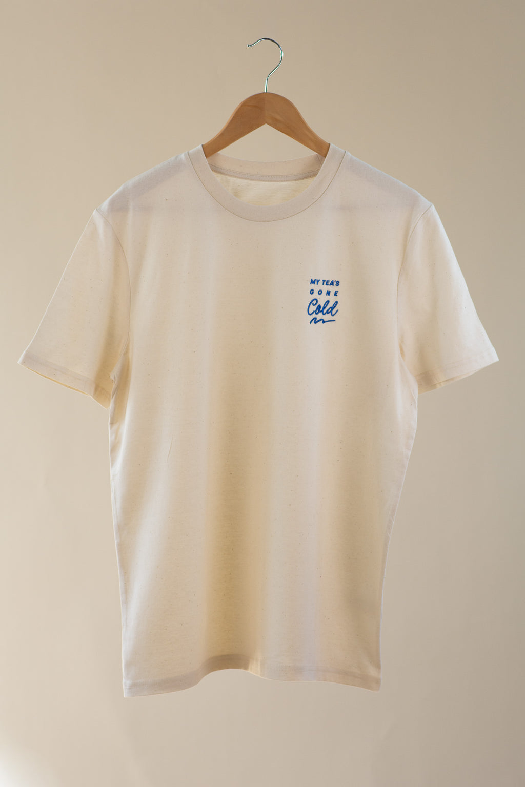 Beige t-shirt with a small blue logo on a wooden hanger against a beige background