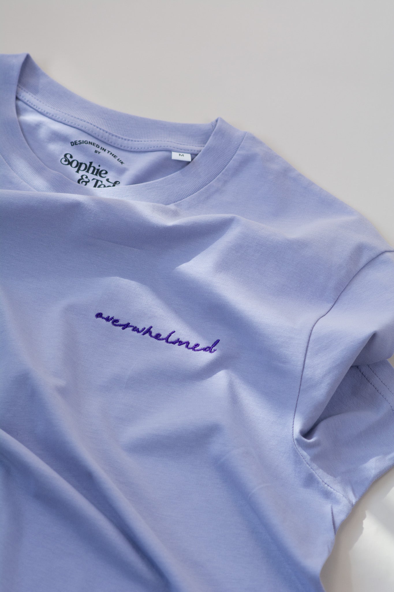 Light purple t-shirt with 'overwhelmed' text and brand logo on a light gray background