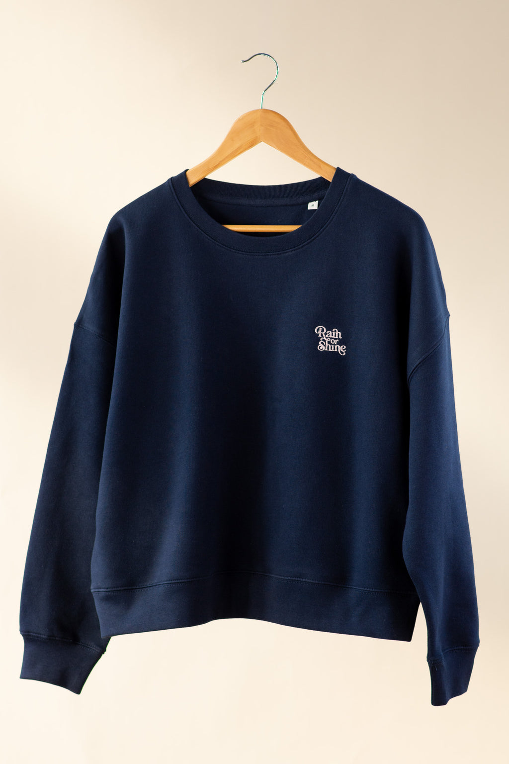 Navy blue sweatshirt with a logo on a wooden hanger against a beige background