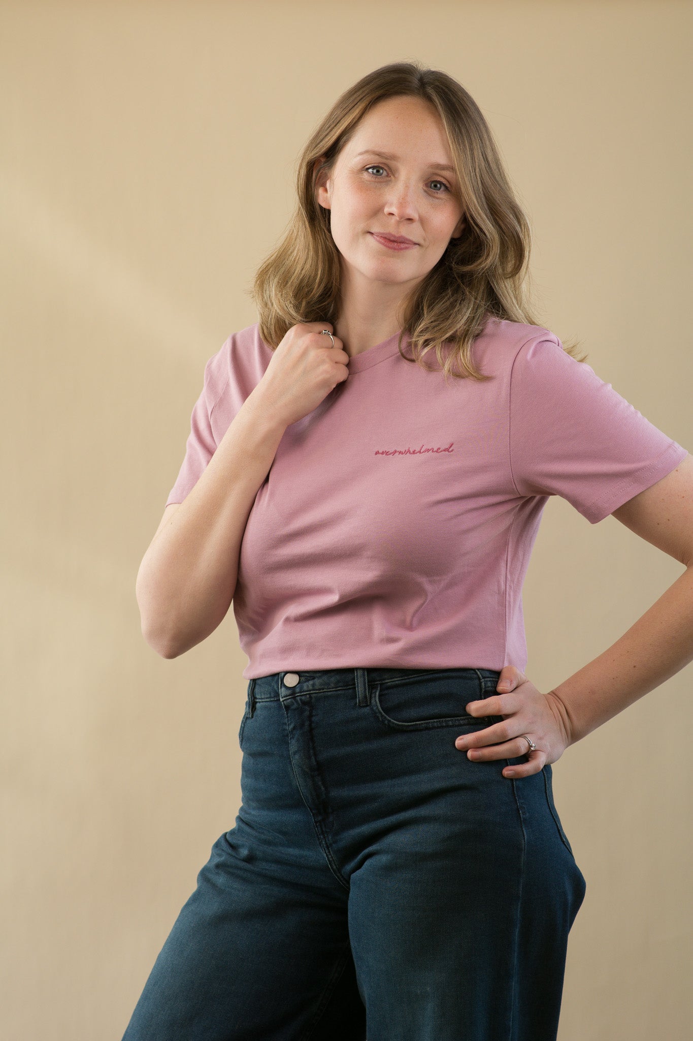 Woman wearing a pink t-shirt with text and dark jeans against a beige background