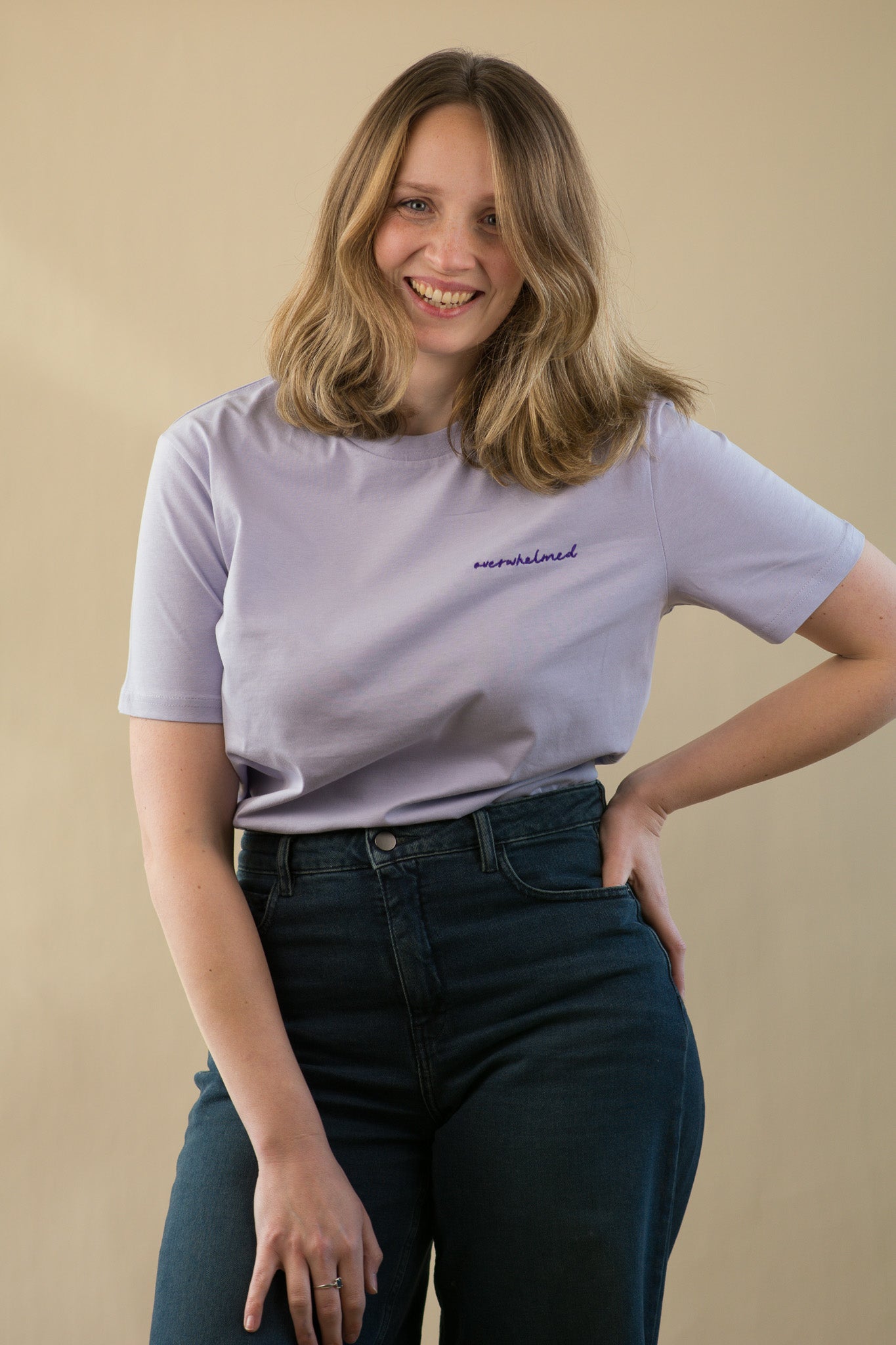 Woman wearing a light purple t-shirt with text and dark jeans against a beige background