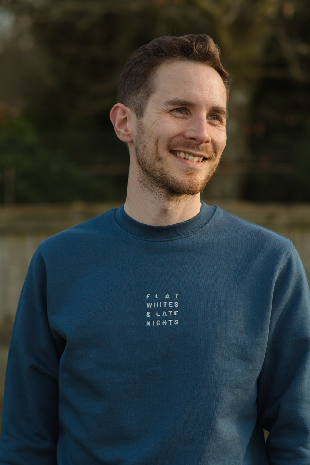 Man wearing a blue sweatshirt with text, standing outdoors with a blurred natural background