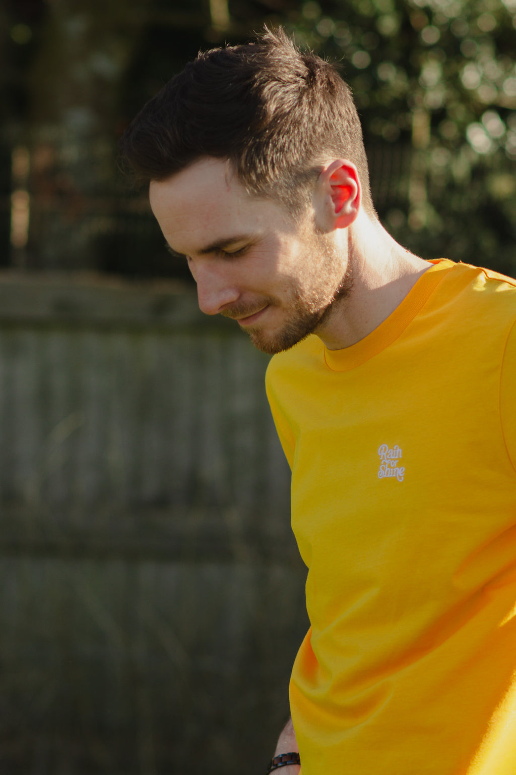 Man wearing a yellow shirt with a logo, standing outdoors with greenery in the background